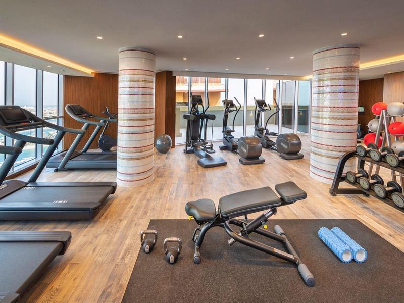 Moderner Fitnessraum mit Laufbändern, Hanteln, Trainingsbank und großen Fenstern mit Stadtblick.