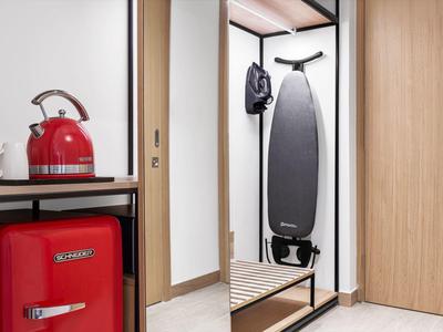 Roter Mini-Kühlschrank und Wasserkocher neben Spiegel mit Bügelbrett und Jacke an der Wand.