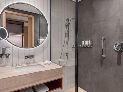 Modernes Badezimmer mit rundem Spiegel, Holzwaschbecken, Glasdusche und grauen Fliesen.