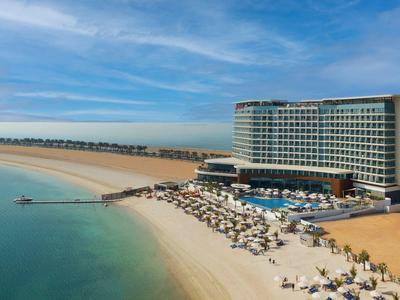 Großes Hotel am Sandstrand mit Pool, Sonnenschirmen und ruhigem Meer unter blauem Himmel.