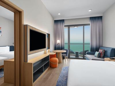 Modernes Hotelzimmer mit zwei Betten, Fernseher, Schreibtisch, Sitzbereich und Meeresblick durch Fenster.