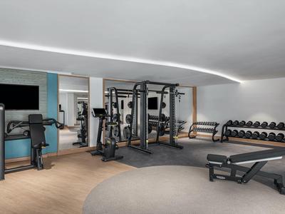 Moderner Fitnessraum mit Kraftgeräten, Hanteln, Spiegeln, hellen Wänden und Holz-Laminatboden.