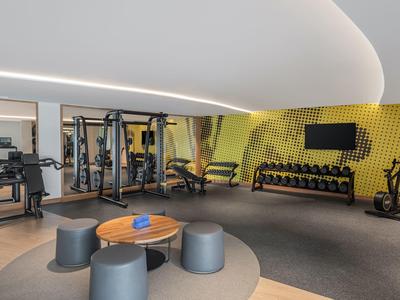 Moderner Fitnessraum mit Geräten, Hanteln, Matten, runden Sitzhockern und blau-gelben Akzenten.
