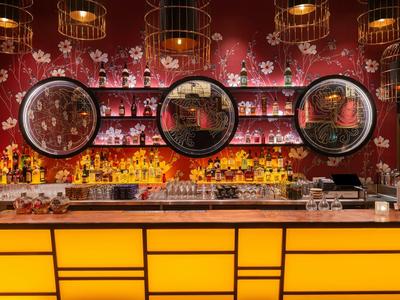 Bar mit gelber Theke, rotem Blumenmuster an der Wand und drei großen Spiegeln über Regalen.