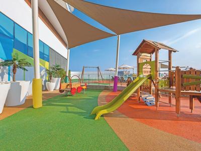Bunter Spielplatz mit Klettergerüst, Rutsche und Schaukeln auf grünem und rotem Bodenbelag.