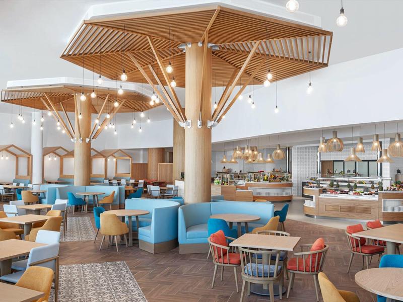 Modernes Restaurant mit hellen Holzmöbeln, bunten Stühlen und Baumstützen mit Beleuchtung.