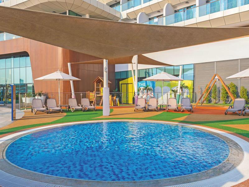 Runder Pool mit Liegestühlen, Sonnenschirmen und modernem Hotelgebäude im Hintergrund.