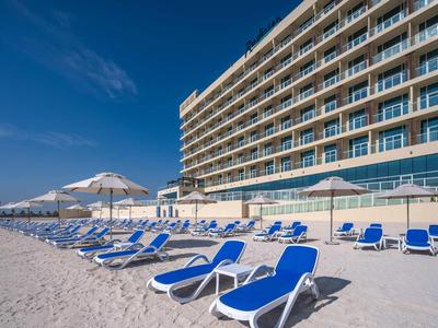 Strandhotel mit blauen Liegestühlen und weißen Sonnenschirmen am Sandstrand unter klarem Himmel.