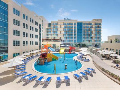 Moderner Außenpool mit Sonnenschirmen, Liegestühlen und Wasserspielplatz in urbaner Hotelumgebung.