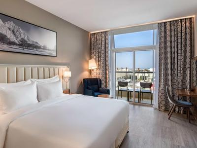 Helles Hotelzimmer mit großem Bett, Stuhl, Schreibtisch und Balkon mit Stadtblick.