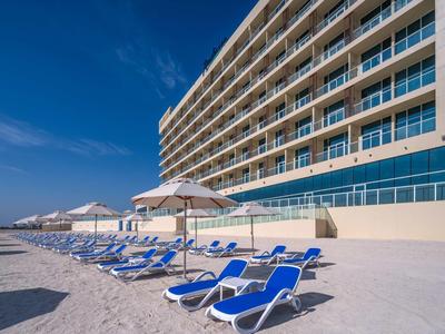 Großes Hotel am Strand mit blauen Liegestühlen und Sonnenschirmen unter klarem Himmel.