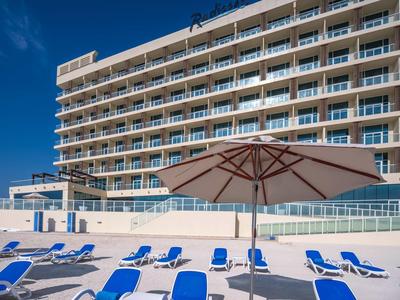 Großes Hotel mit Balkonen, Sonnenschirmen und blauen Liegestühlen am Strand unter blauem Himmel.