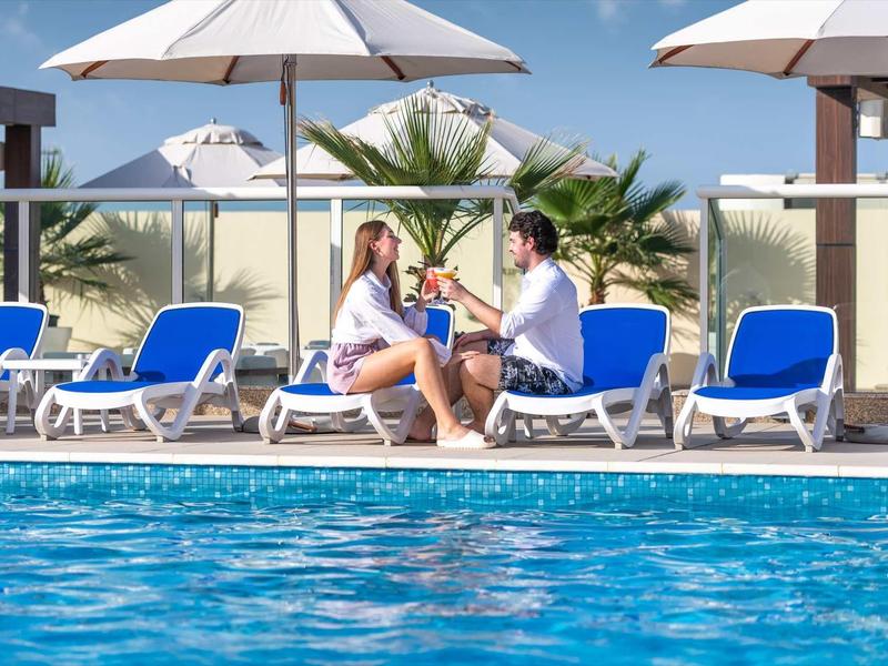 Paar sitzt am Pool auf Liegestühlen, trinkt Cocktails, viele blaue Liegen und Sonnenschirme.