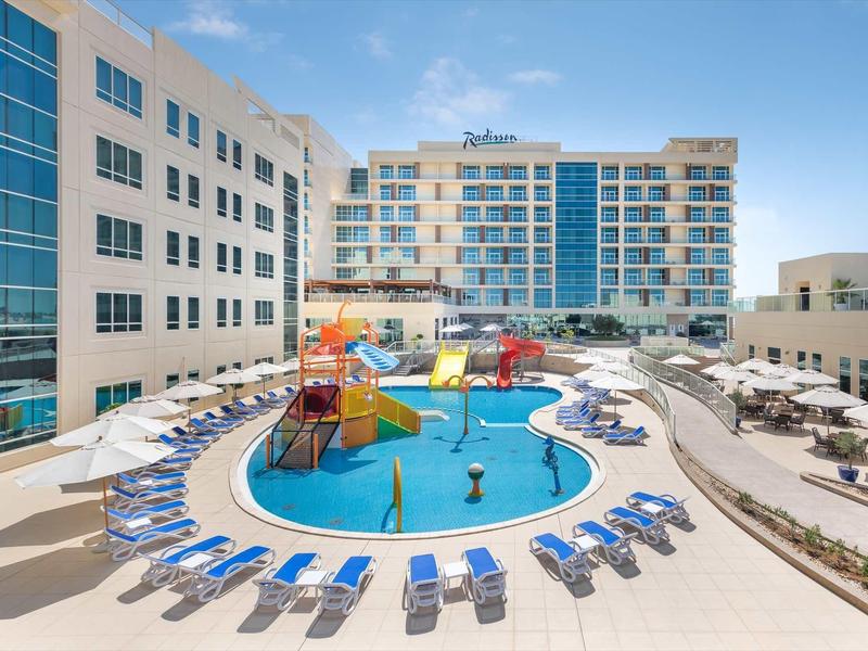 Moderner Außenpool mit Sonnenschirmen, Liegestühlen und Wasserspielplatz in urbaner Hotelumgebung.