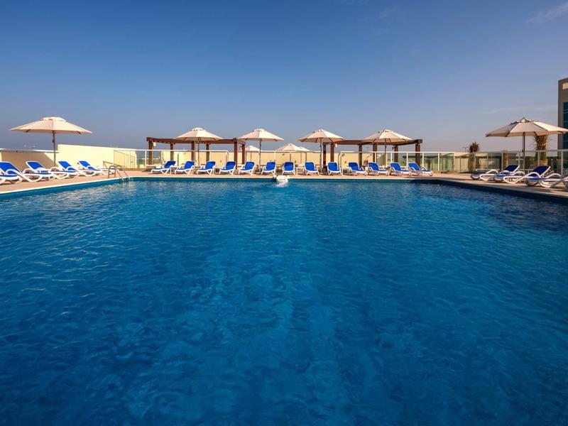 Großes blaues Schwimmbecken mit Liegestühlen und Sonnenschirmen unter klarem Himmel.