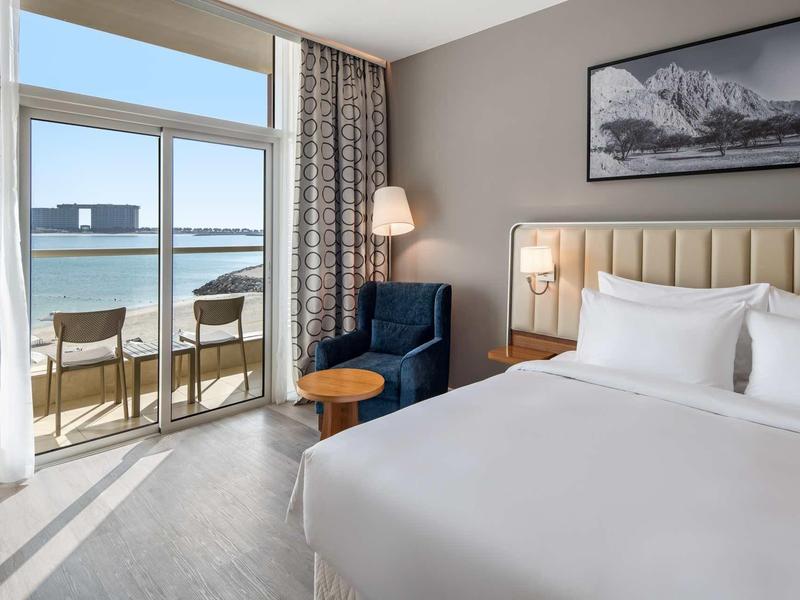 Modernes Hotelzimmer mit großem Bett, Balkon und Meerblick, helles Farbenkonzept, gemütlicher Sessel.