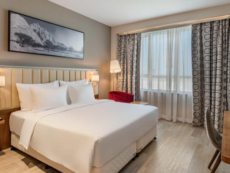 Modern eingerichtetes Hotelzimmer mit großem Bett, Nachttischen, Vorhängen und Fensterfront.