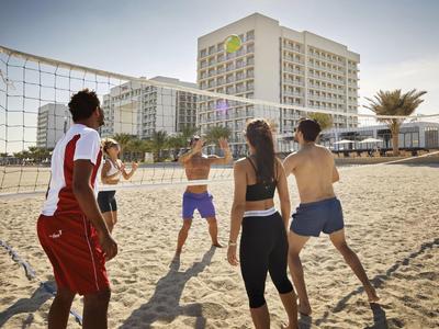 Fünf Menschen spielen Volleyball am Strand vor großen Hotels unter blauem Himmel.