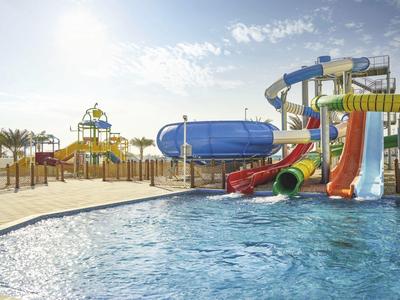 Wasserpark mit bunten Wasserrutschen, Becken und Sonnenschein unter blauem Himmel.