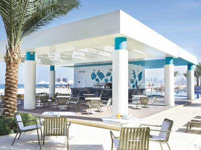 Moderner Pavillon mit weißen Säulen, blauen Akzenten und Sitzgelegenheiten in sonniger Umgebung.