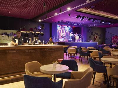 Moderner Club mit Bühne, Stühlen und Tischen, lila Beleuchtung, Sänger auf der Bühne, ruhige Atmosphäre.