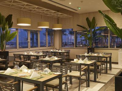 Modernes Restaurant mit dunklen Tischen, vielen Stühlen und grünen Zimmerpflanzen in großen Fenstern.