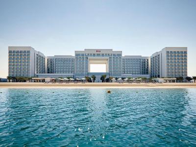 Modernes Hotel mit weißer Fassade, großem Mittelteil und Meer im Vordergrund bei klarem Himmel.