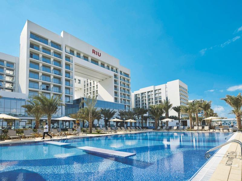 Modern hotel with large pool, sun loungers, and palm trees under a blue sky.
