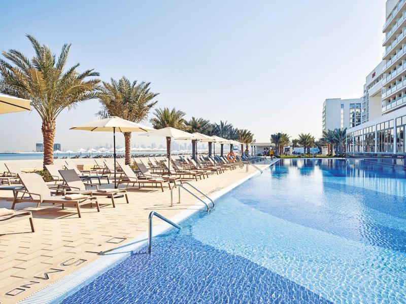 Modern hotel pool with loungers, umbrellas, and palm trees under a clear sky.