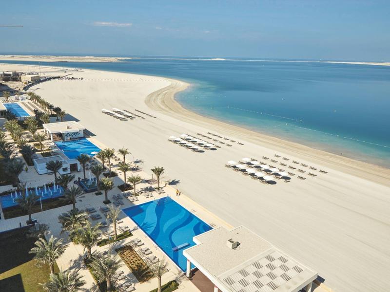 Wide sandy beach with sun loungers and blue hotel pool beside calm sea