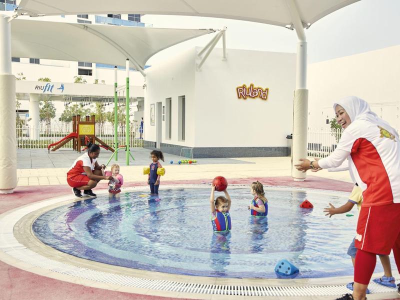 Children play in the shallow pool under shade with supervisors in red and white clothing.