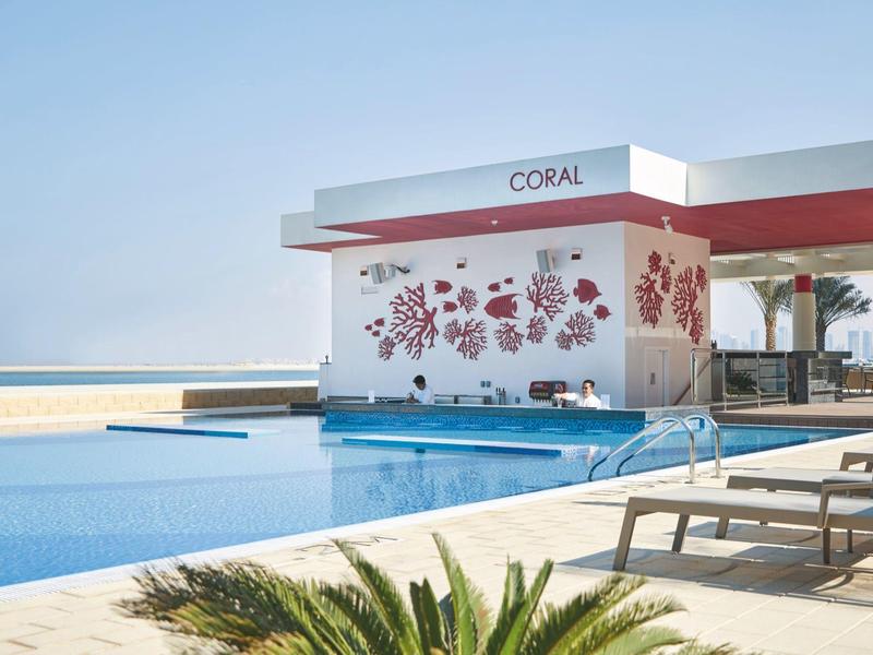 Modern hotel pool with loungers, bar, and sea view under clear sky