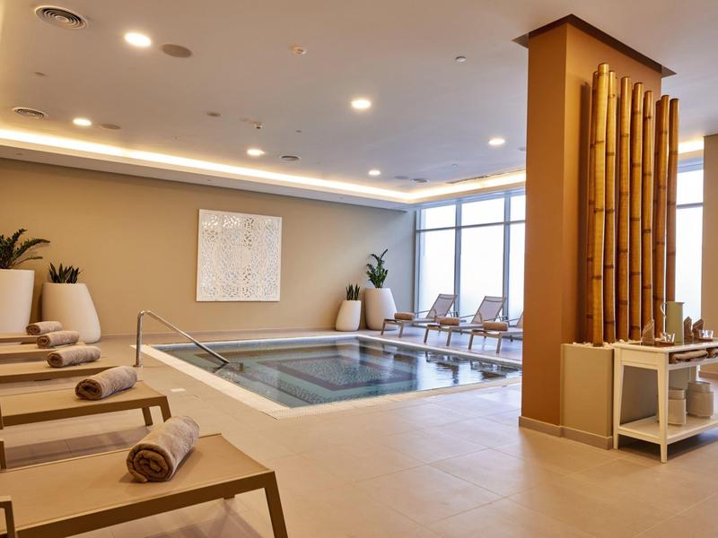 Modern indoor pool area with lounge chairs, plants, and natural light from large windows.