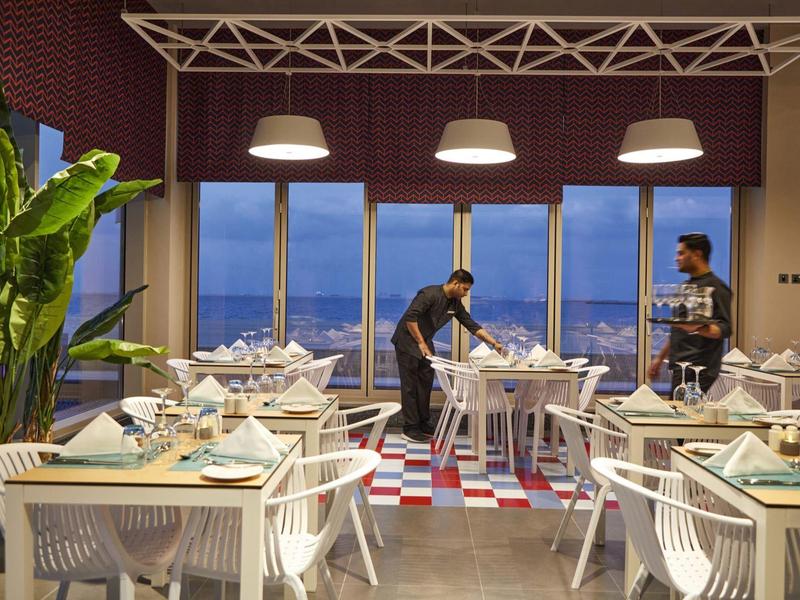 Modern restaurant with white chairs, large plants, and sea view at sunset.