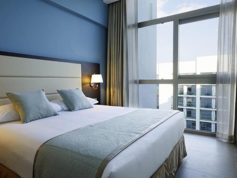 Modern hotel room with large bed, gray pillows, and floor-to-ceiling windows overlooking the city.