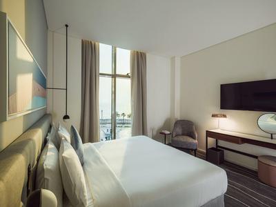 Modernes Hotelzimmer mit großem Bett, Fernseher, Sessel und bodentiefem Fenster mit Blick nach draußen.
