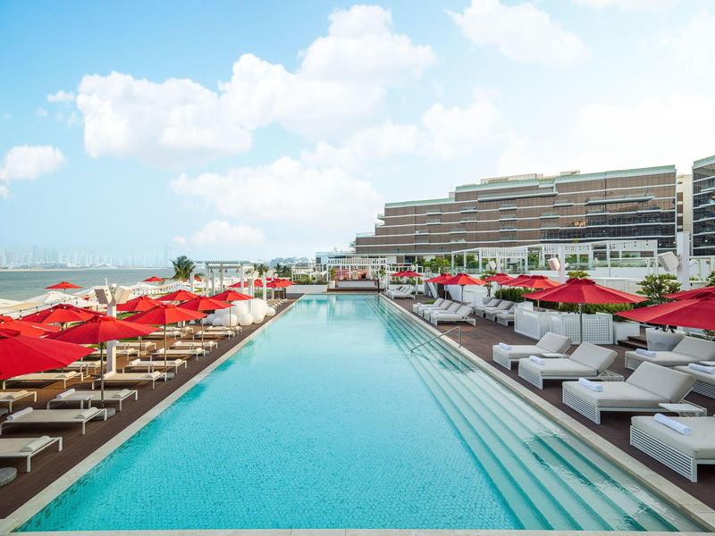 Schwimmbad mit klarem Wasser, umgeben von roten Sonnenschirmen und weißen Liegen, neben modernen Gebäuden.