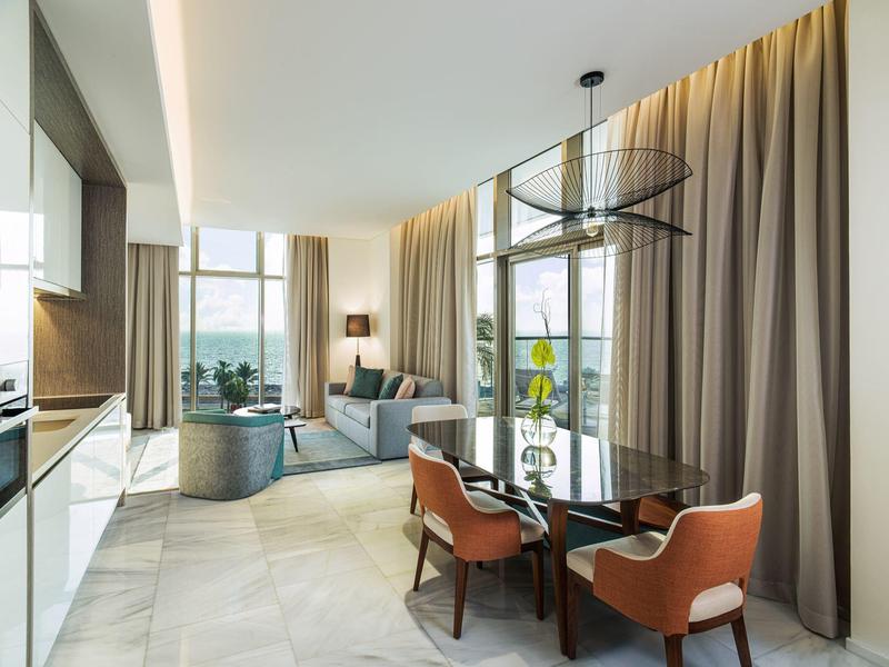 Heller moderner Wohnraum mit Glas-Tisch, orangen Stühlen, grauer Couch und großen Fenstern.