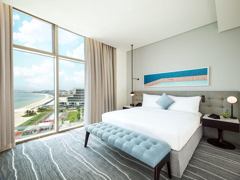 Modernes Hotelzimmer mit großem Bett, blauer Bank und Meerblick durch große Fenster.