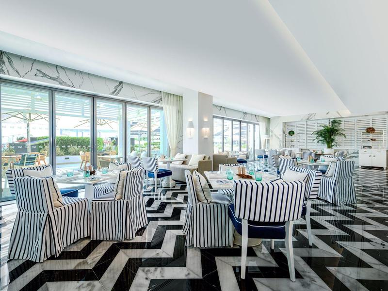 Heller Hotel-Wintergarten mit gestreiften Sesseln und großflächigen Fenstern zum Außenbereich.