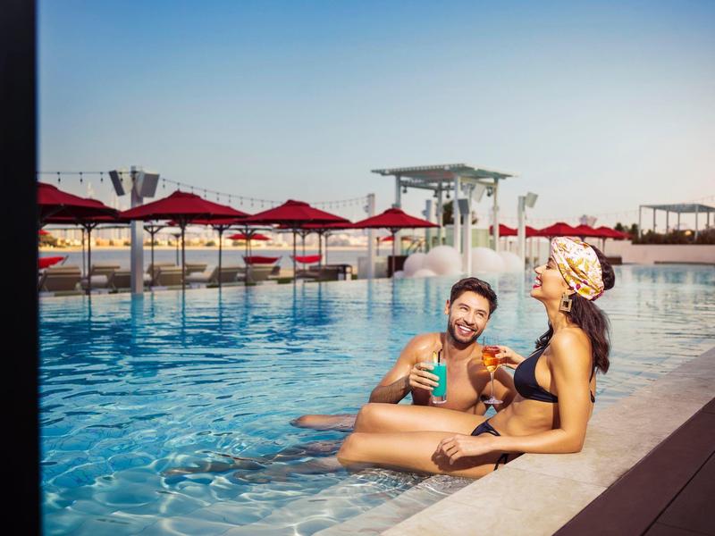 Zwei Menschen sitzen am Poolrand, genießen bunte Drinks bei sonnigem Wetter und roten Sonnenschirmen.
