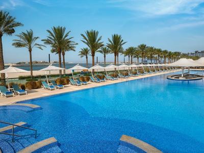 Una piscina larga con tumbonas y sombrillas, rodeada de palmeras bajo un cielo azul.
