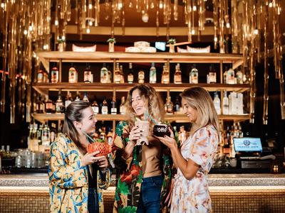 Tres mujeres riendo y sosteniendo cócteles en un bar con ambiente festivo.