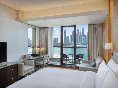 Modernes Hotelzimmer mit großem Bett, TV, zwei Sesseln und großem Fenster mit Blick auf Stadt und Wasser.