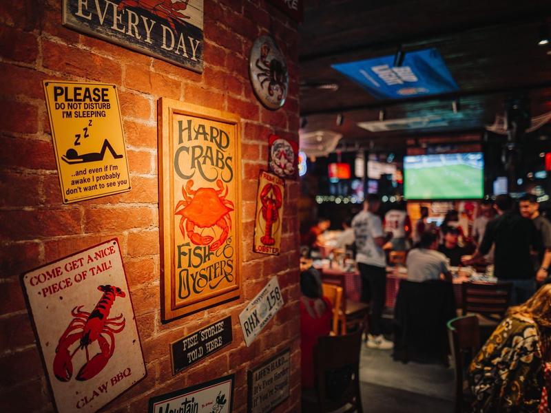 Gemütliche Bar mit Backsteinwand und bunten Schildern, im Hintergrund Gäste und Fernseher.