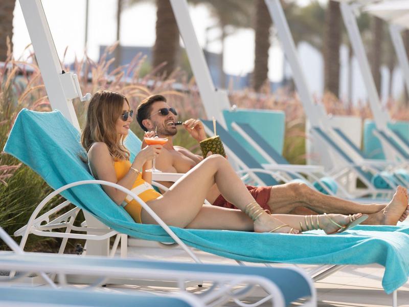Zwei Personen entspannen auf Liegestühlen am Strand unter Sonnenschirmen.