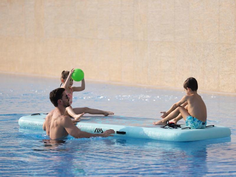 Zwei Kinder spielen mit einem Eimer auf einer blauen Luftmatratze im Pool.