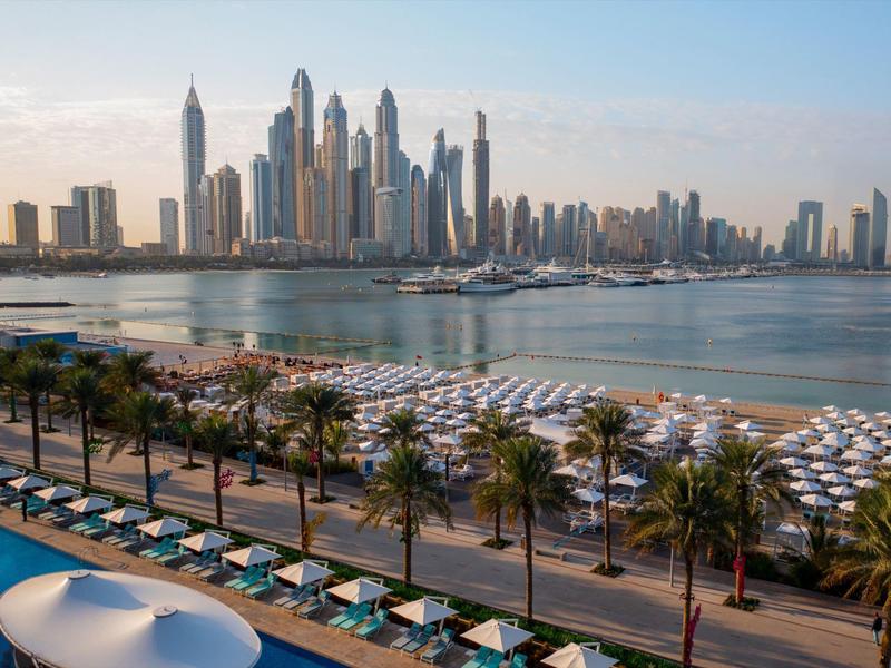 Stadt mit vielen Wolkenkratzern, Strand mit Schirmen, Palmen und blauem Wasser im Vordergrund.