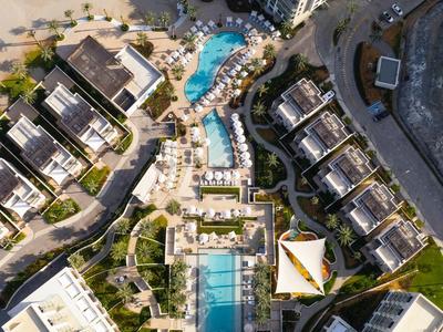 Luftaufnahme einer Hotelanlage mit mehreren Pools, Sonnenschirmen und angrenzenden Gebäuden am Strand.