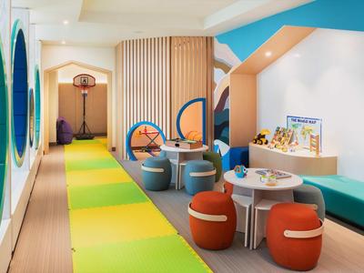 Heller Kinderspielraum mit buntem Boden, Sitzhockern, Basketballkorb und Spielzeugregal entlang der Wand.